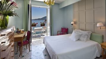 A hotel room with a bed, a desk with a chair, a dresser, a balcony with a table and chairs, and a view of the sea.