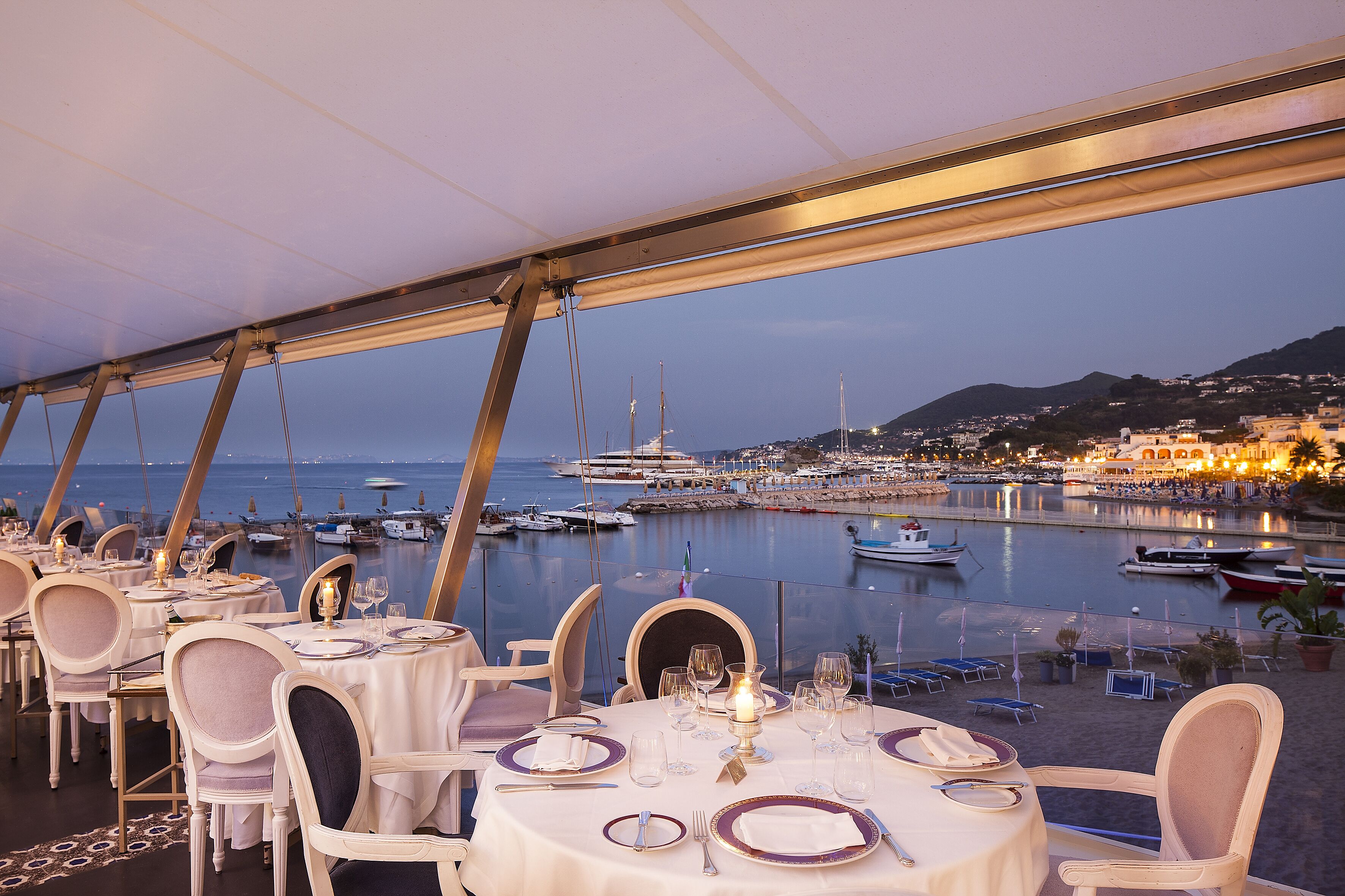 A restaurant with tables set for dining, overlooking a marina with boats and a cityscape in the background.