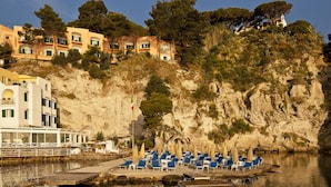 Un hôtel en bord de mer doté d’une terrasse offrant une vue sur l’océan, avec une rangée de chaises longues bleues et une falaise rocheuse en arrière-plan.