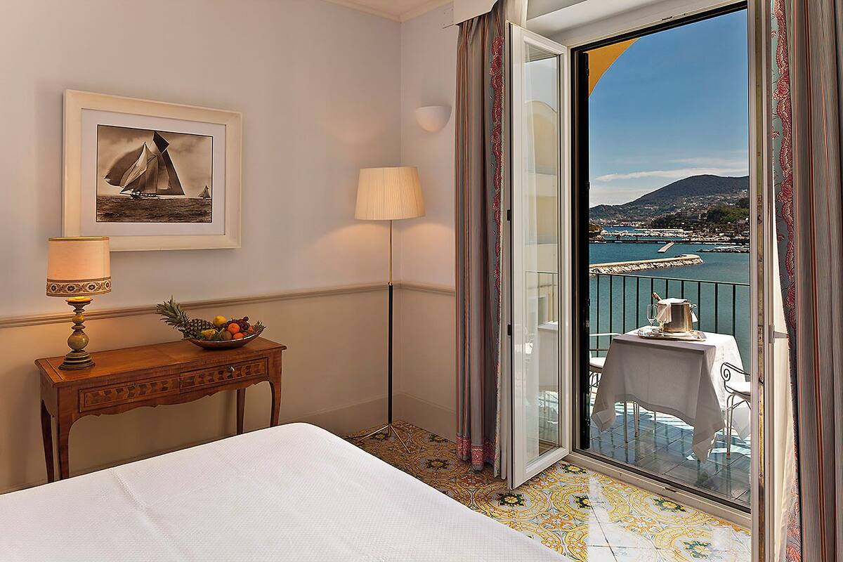 Une chambre d’hôtel avec un lit, une petite table sur laquelle se trouvent une lampe et un panier de fruits, un tableau encadré, et un balcon avec une table préparée pour deux personnes, offrant une vue sur la côte.
