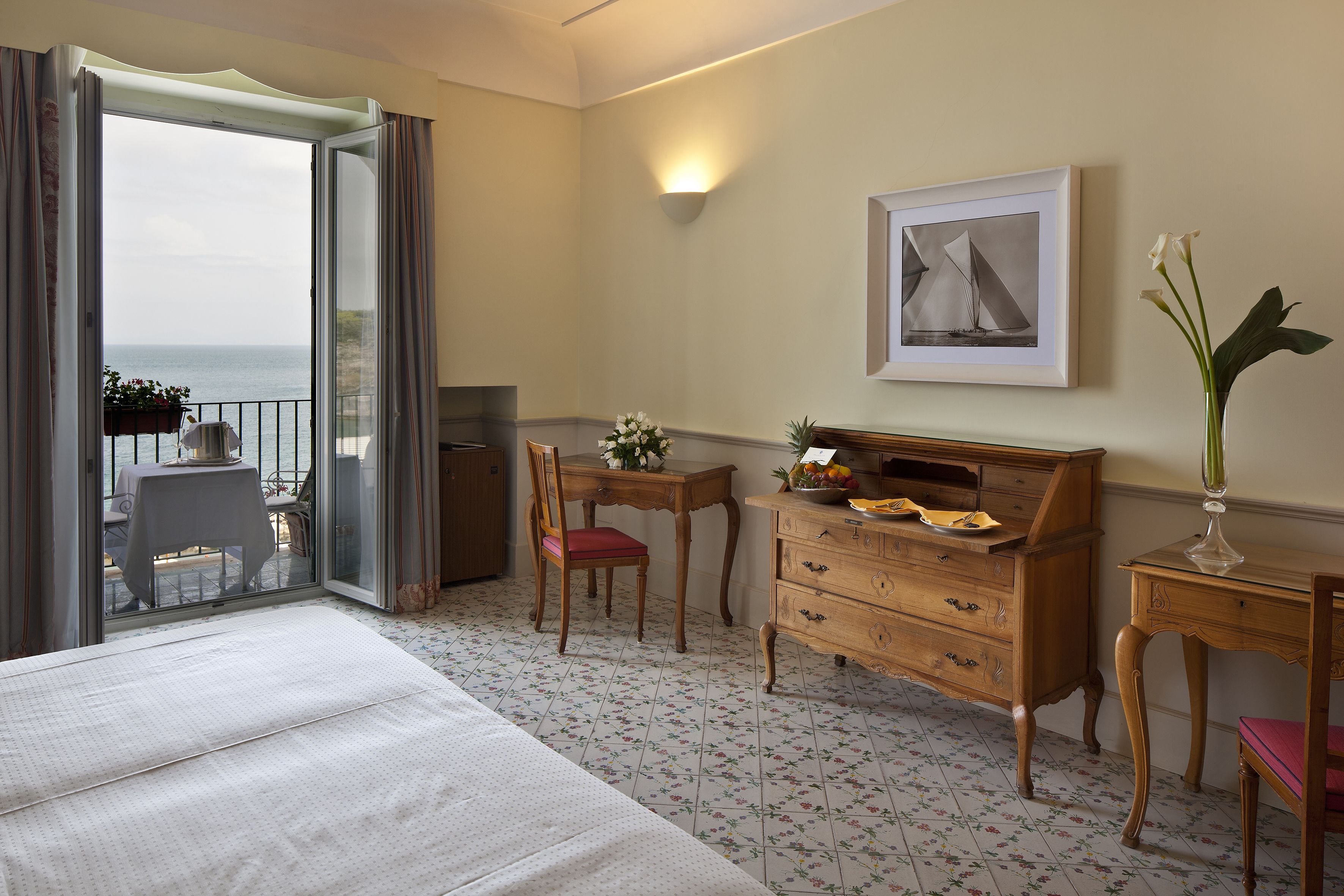 A hotel room with a bed, a desk, a chair, a dresser, a potted plant, a framed picture, and a view of the sea through a balcony door.
