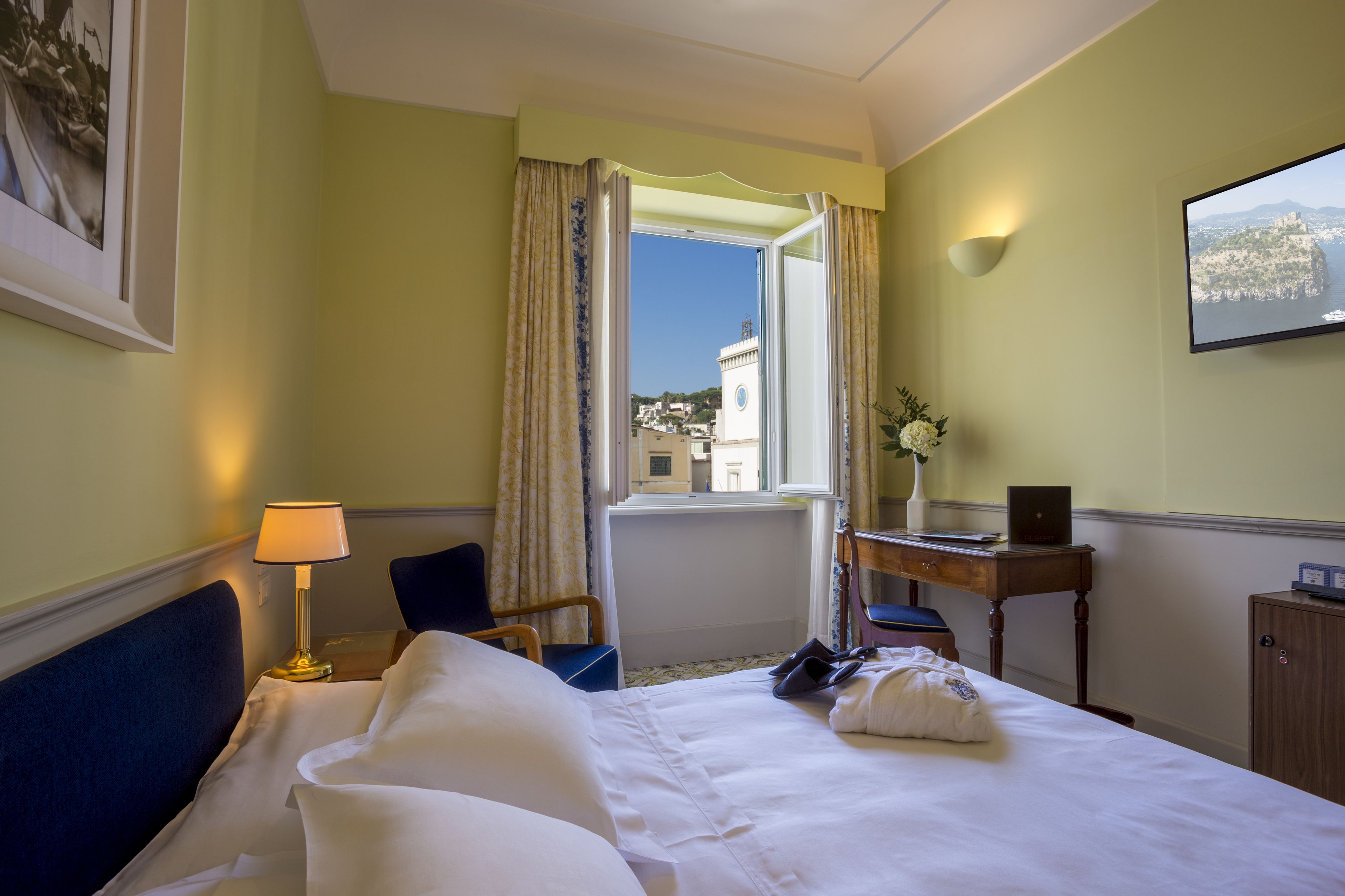A hotel room with a bed, a desk with a laptop, a chair, a lamp, and a view of a cityscape through a window.