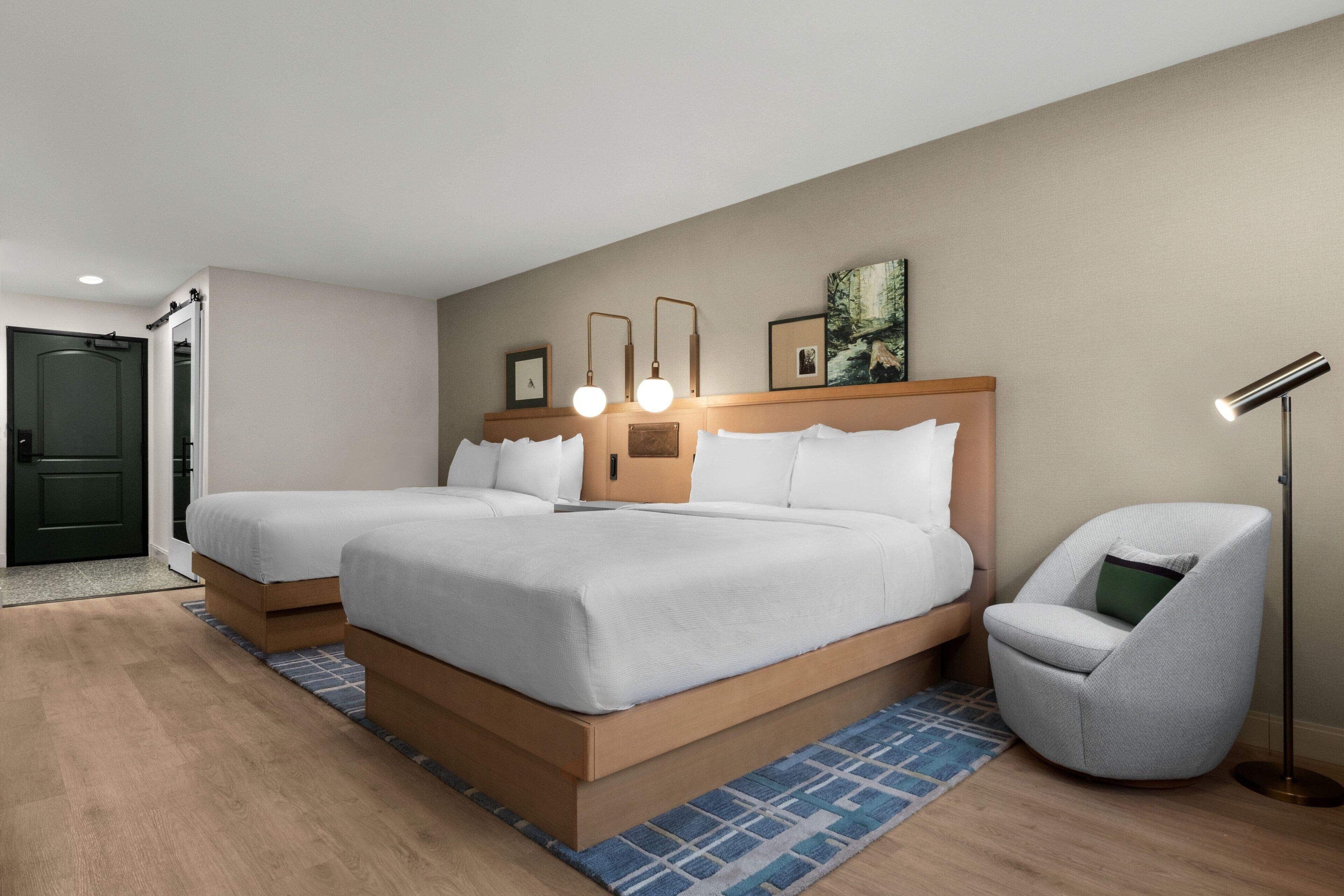 A hotel room with two beds, a chair, a floor lamp, and a door.