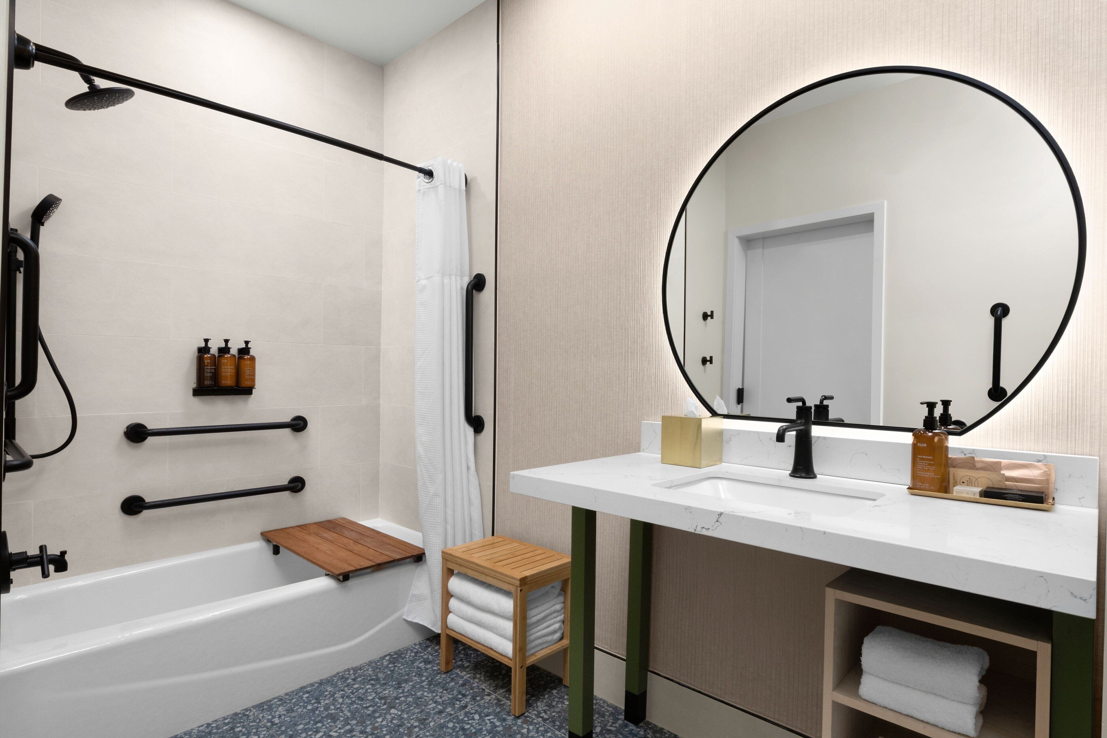 A modern bathroom with a bathtub, a wooden stool, a sink with a mirror, and toiletries.