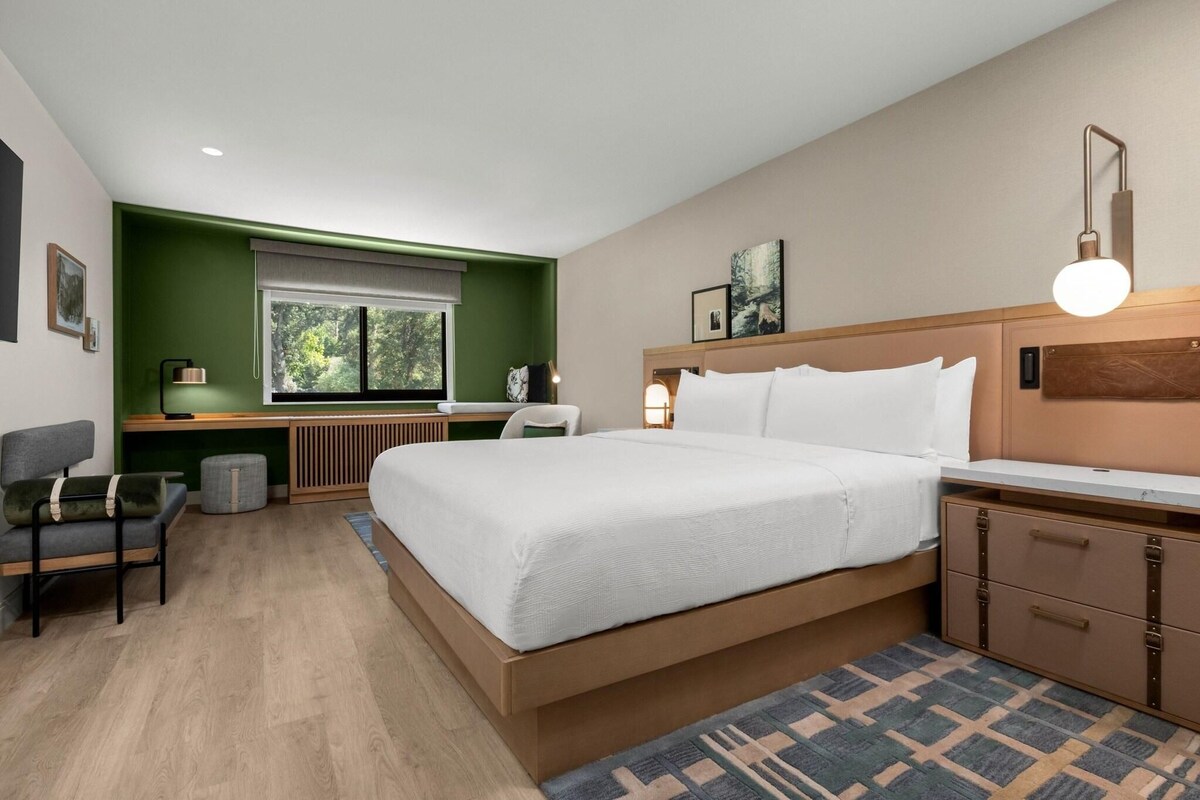 Quarto de hotel moderno com uma cama grande, uma escrivaninha, uma cadeira e uma janela com vista para árvores.