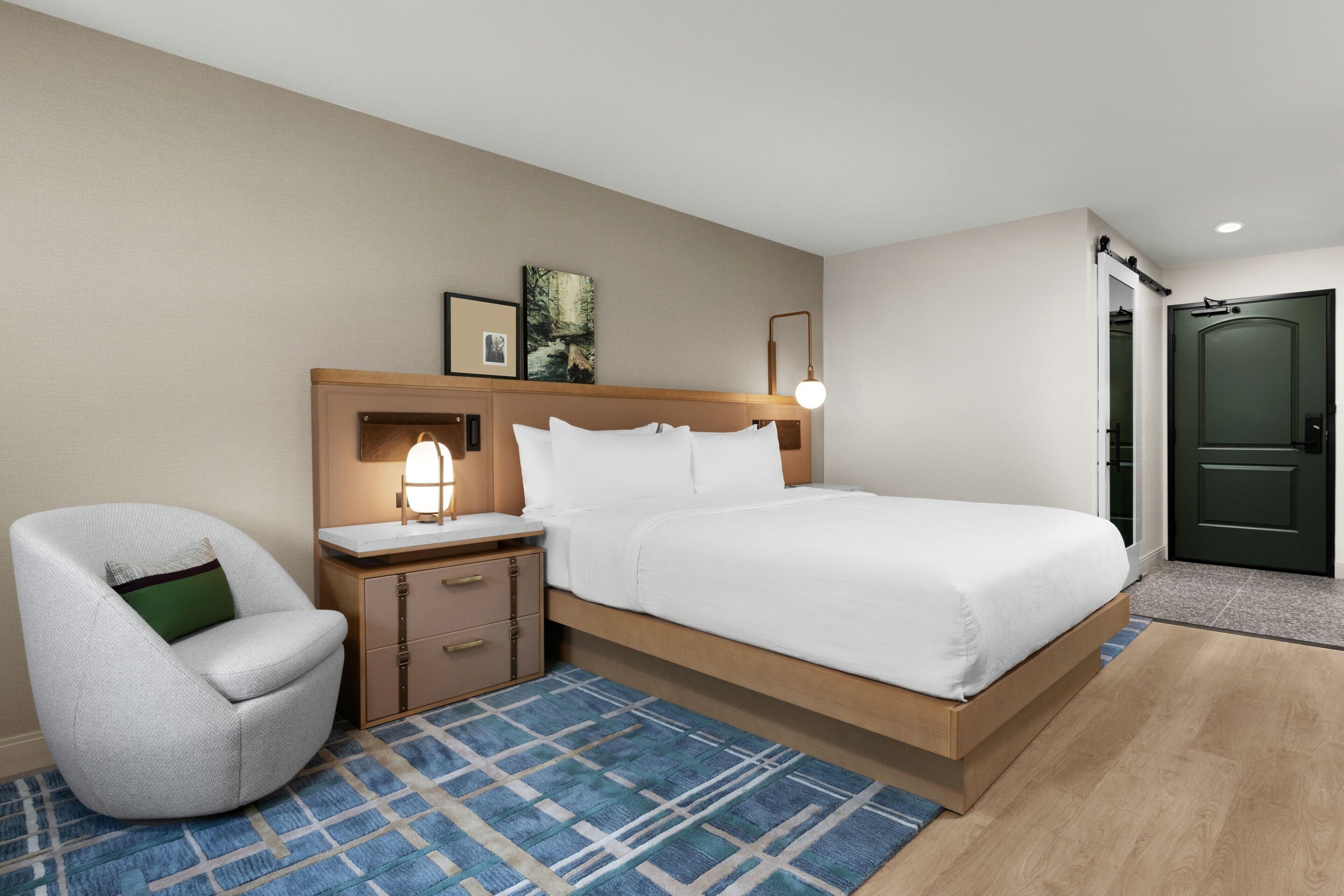 Una habitación de hotel con cama, una silla, una mesita de noche, una lámpara y una puerta.