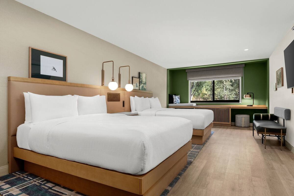 A modern hotel room with two beds, a desk, and a view of the outdoors.