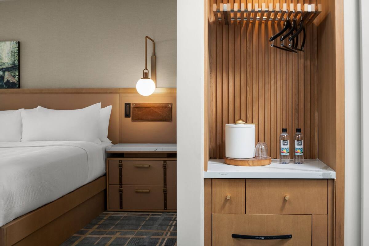 A hotel room with a bed, bedside table, lamp, and wooden panel wall with hooks.