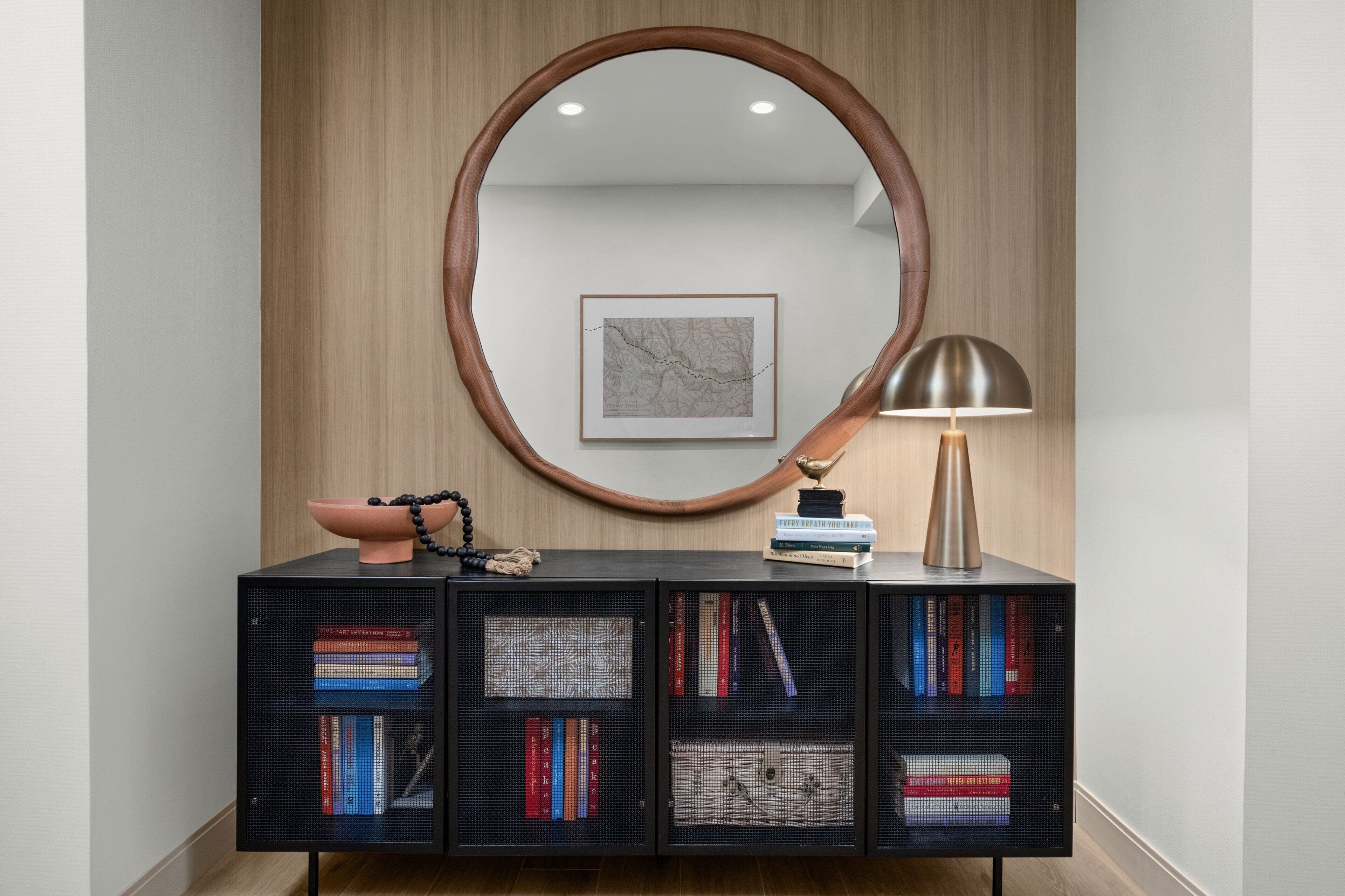 Un interior moderno con un panel de madera, un espejo redondo grande, una consola negra con libros, un cuenco decorativo y una lámpara.