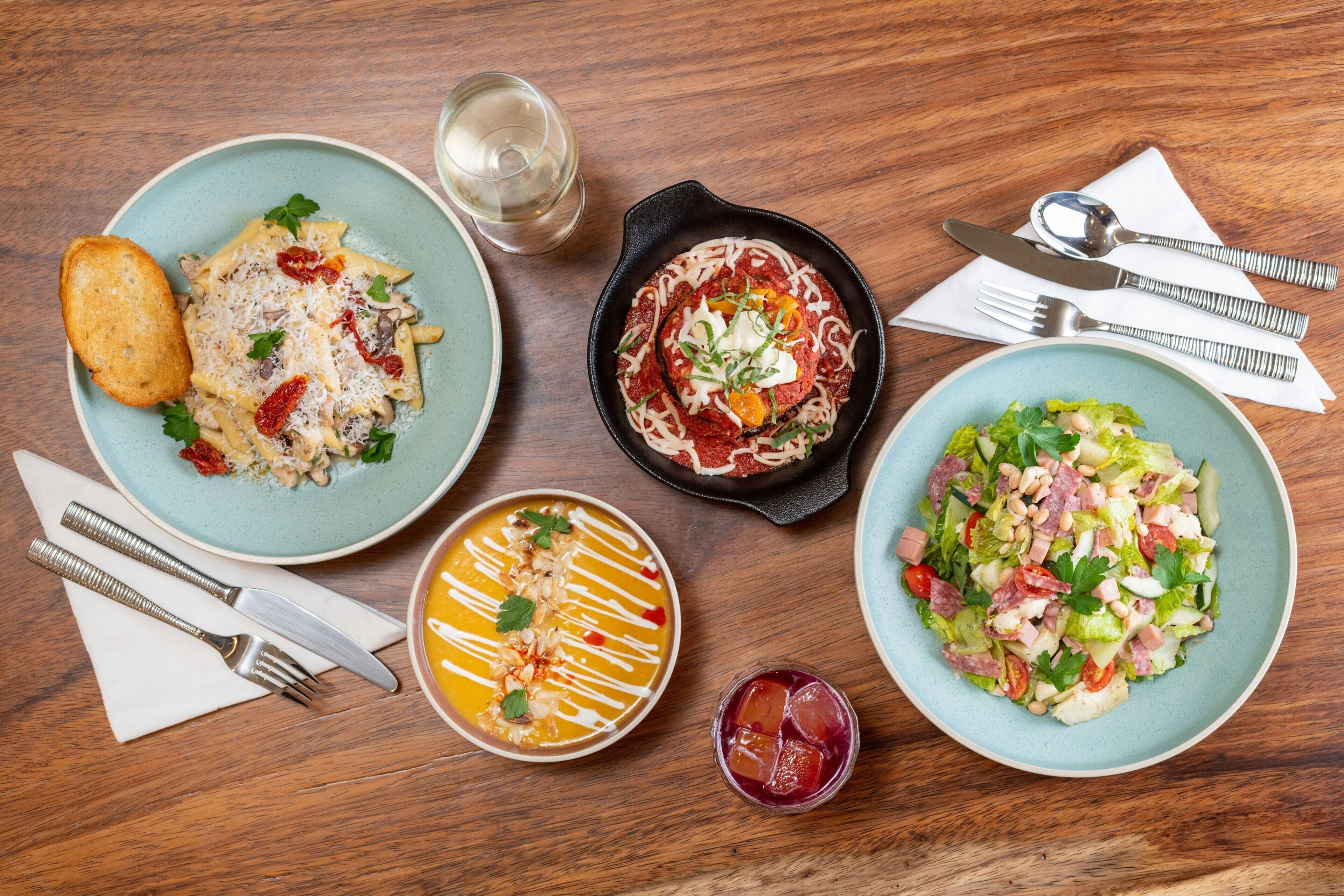 Een houten tafel met diverse gerechten, waaronder pasta, een salade en een soepkom, plus bestek en een glas water.