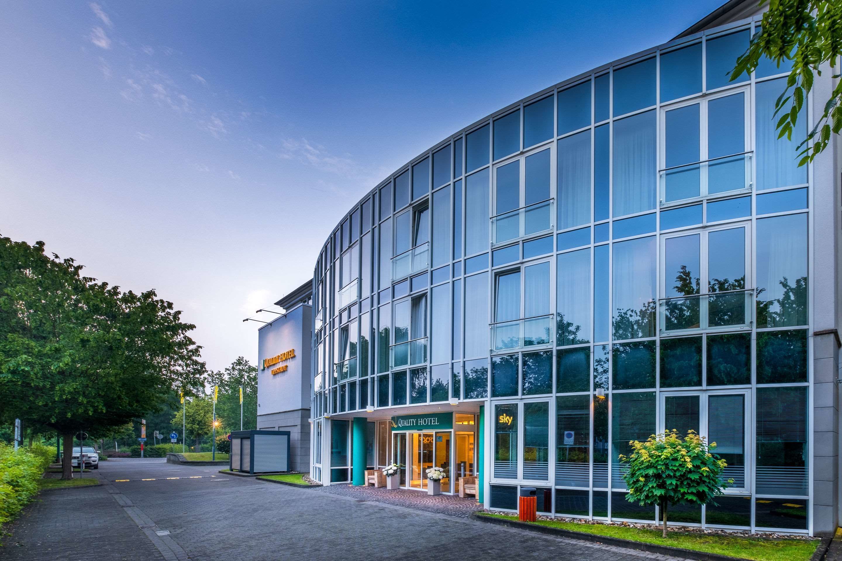 A modern hotel building with a glass facade, a sign for "Quality Hotel," and a well-maintained garden area.