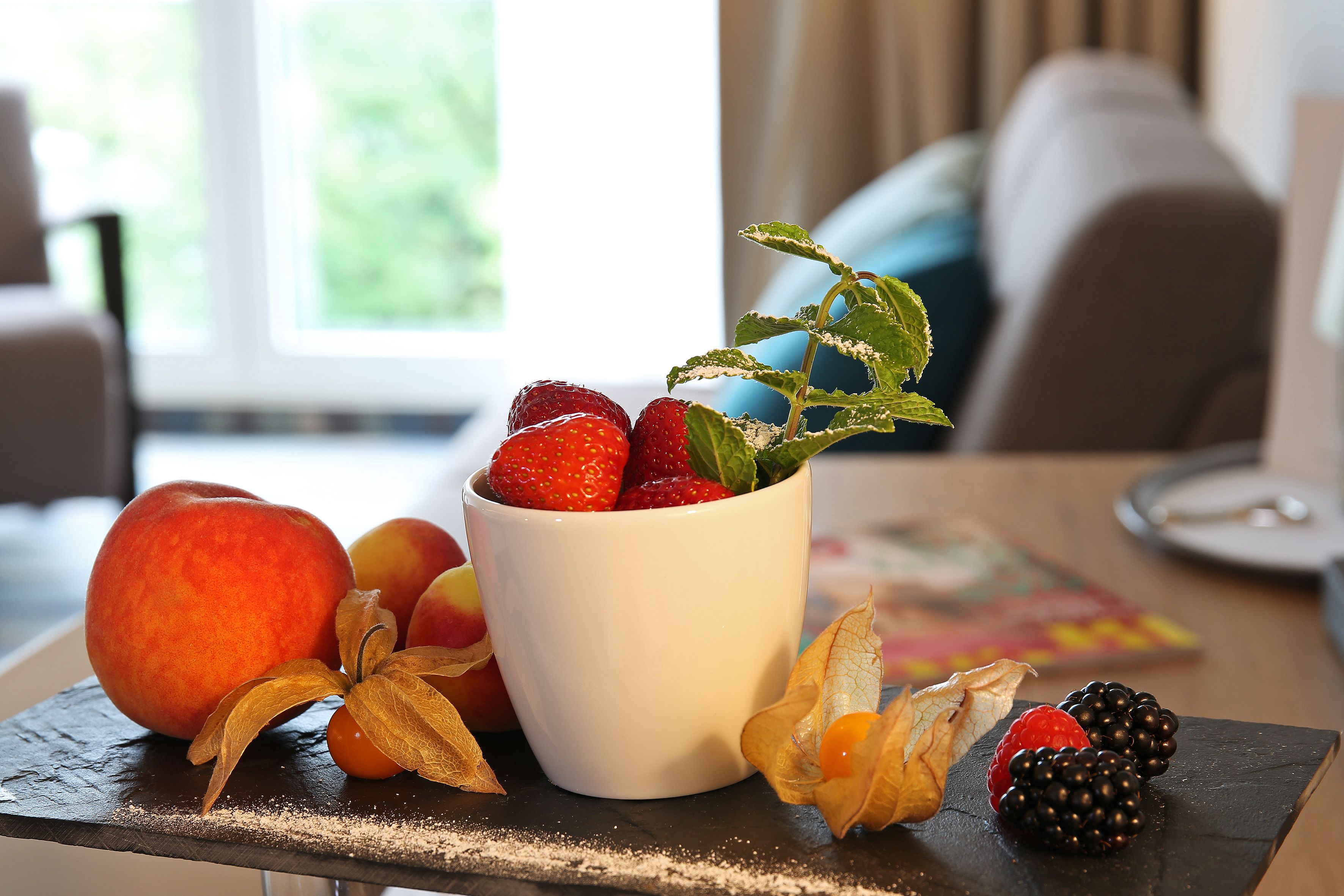 Obstschale auf einem Tisch, mit einer weißen Schüssel, die Erdbeeren, einen Pfirsich und getrocknete Blumen enthält.