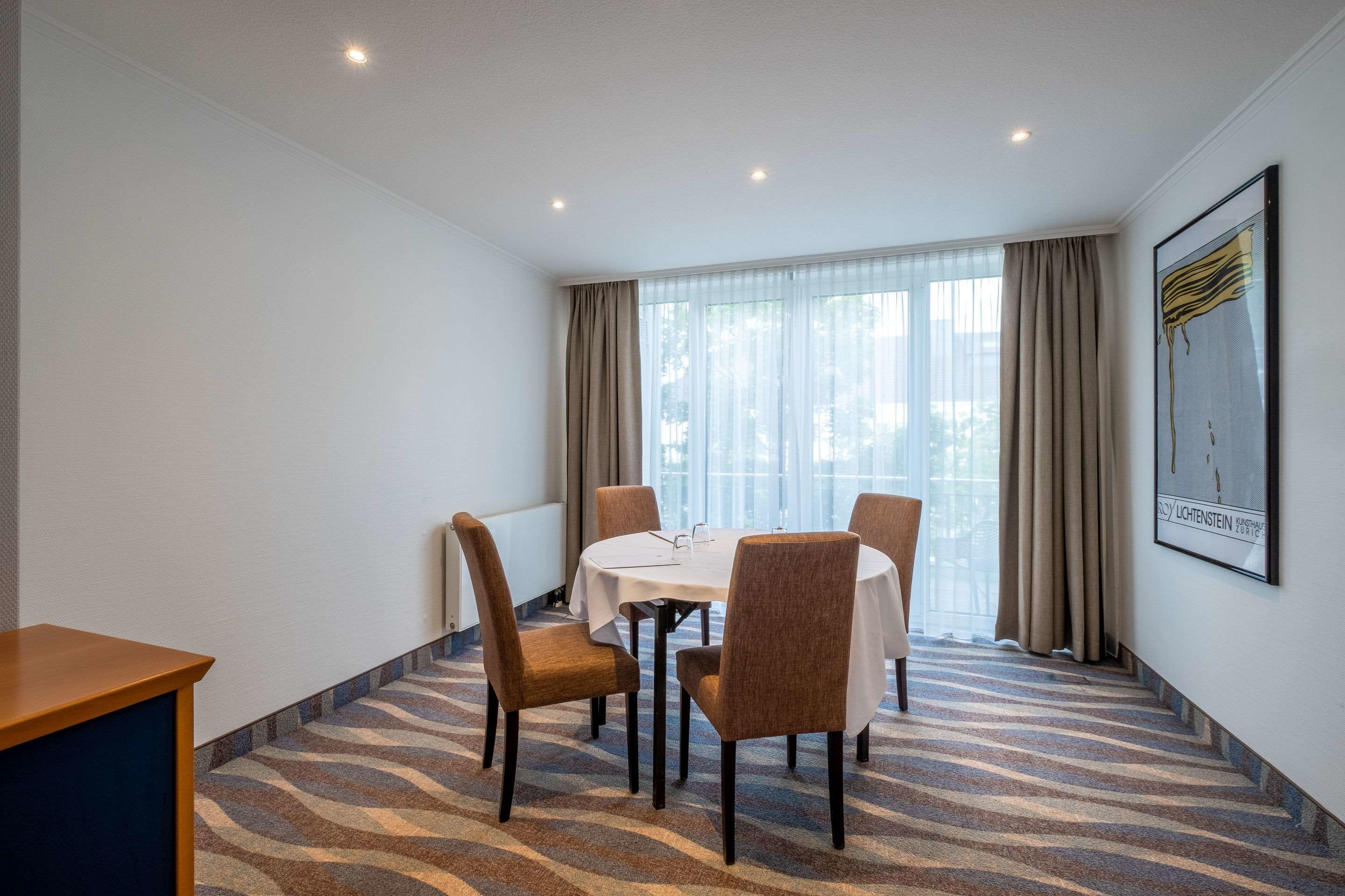 Une chambre d’hôtel avec une table ronde, quatre chaises, une fenêtre avec des rideaux et un tableau encadré au mur.