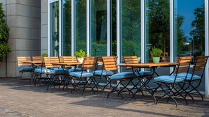 An outdoor seating area with wooden tables and chairs, potted plants, and a view of trees through large windows. - Quality Hotel Lippstadt (Lippstadt)