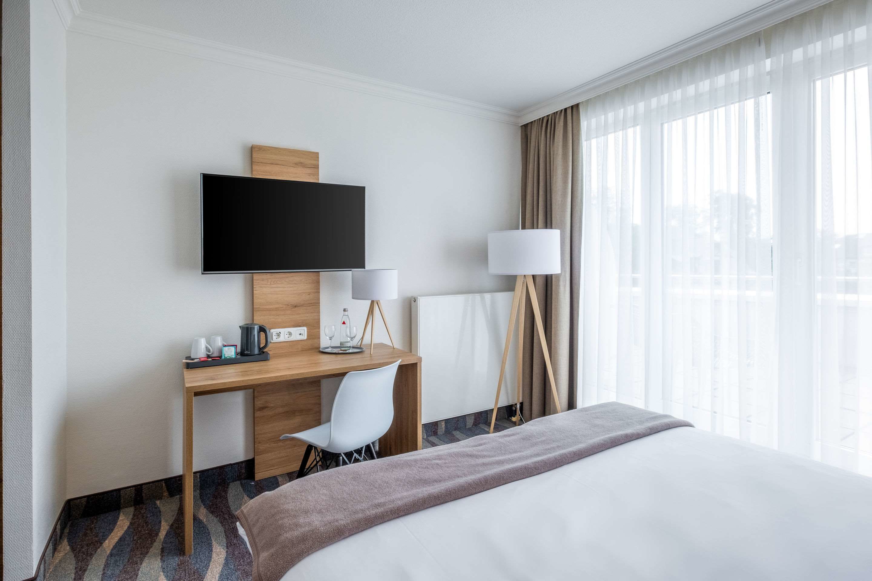 Une chambre d’hôtel comprenant un lit, un bureau avec une chaise, un téléviseur à écran plat fixé au mur, une lampe et une fenêtre avec des rideaux.