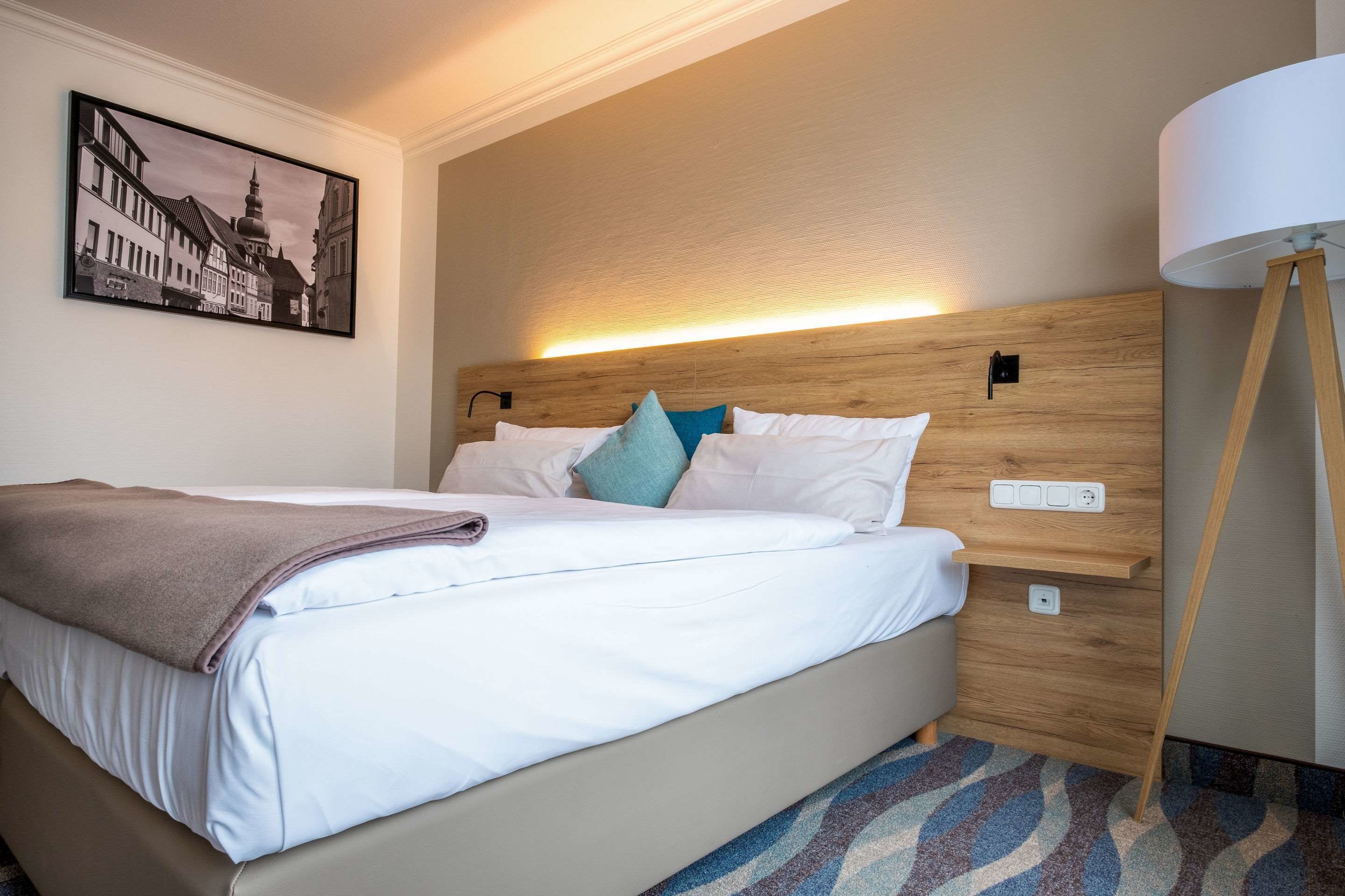 A neatly made bed with a brown blanket, a headboard with a light fixture, a wall-mounted power outlet, and a patterned rug.