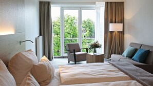 A hotel room with a large bed, a sofa, a chair, a small table with a lamp, and a view of greenery outside. - Quality Hotel Lippstadt (Lippstadt)