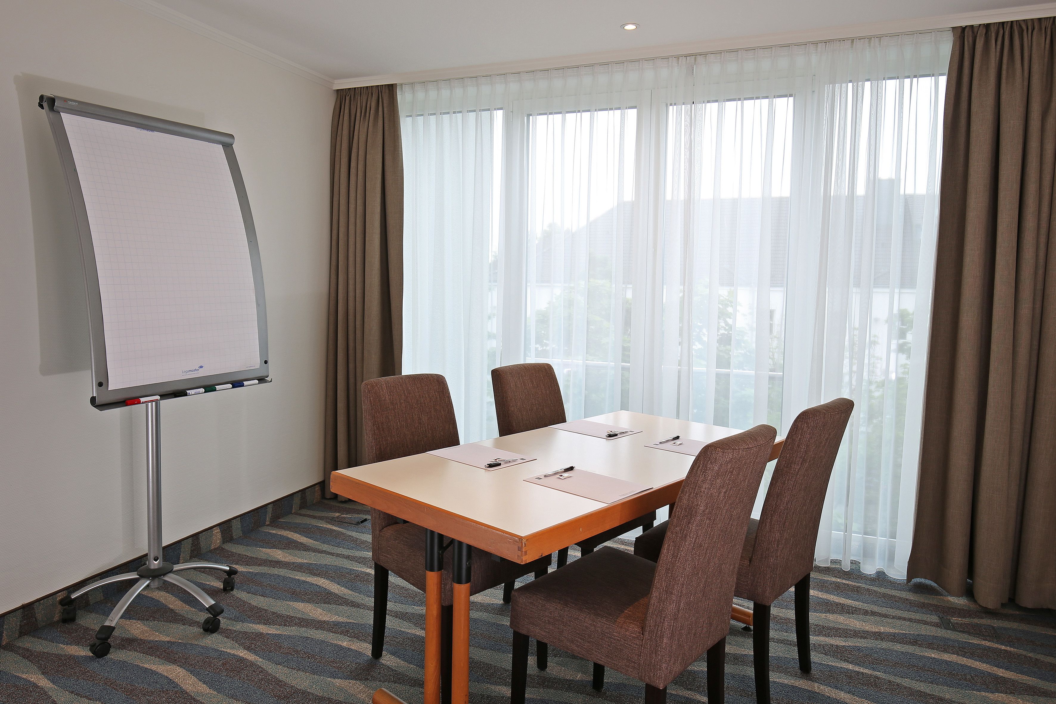 A conference room with a large table, four chairs, a whiteboard, and a window with curtains.