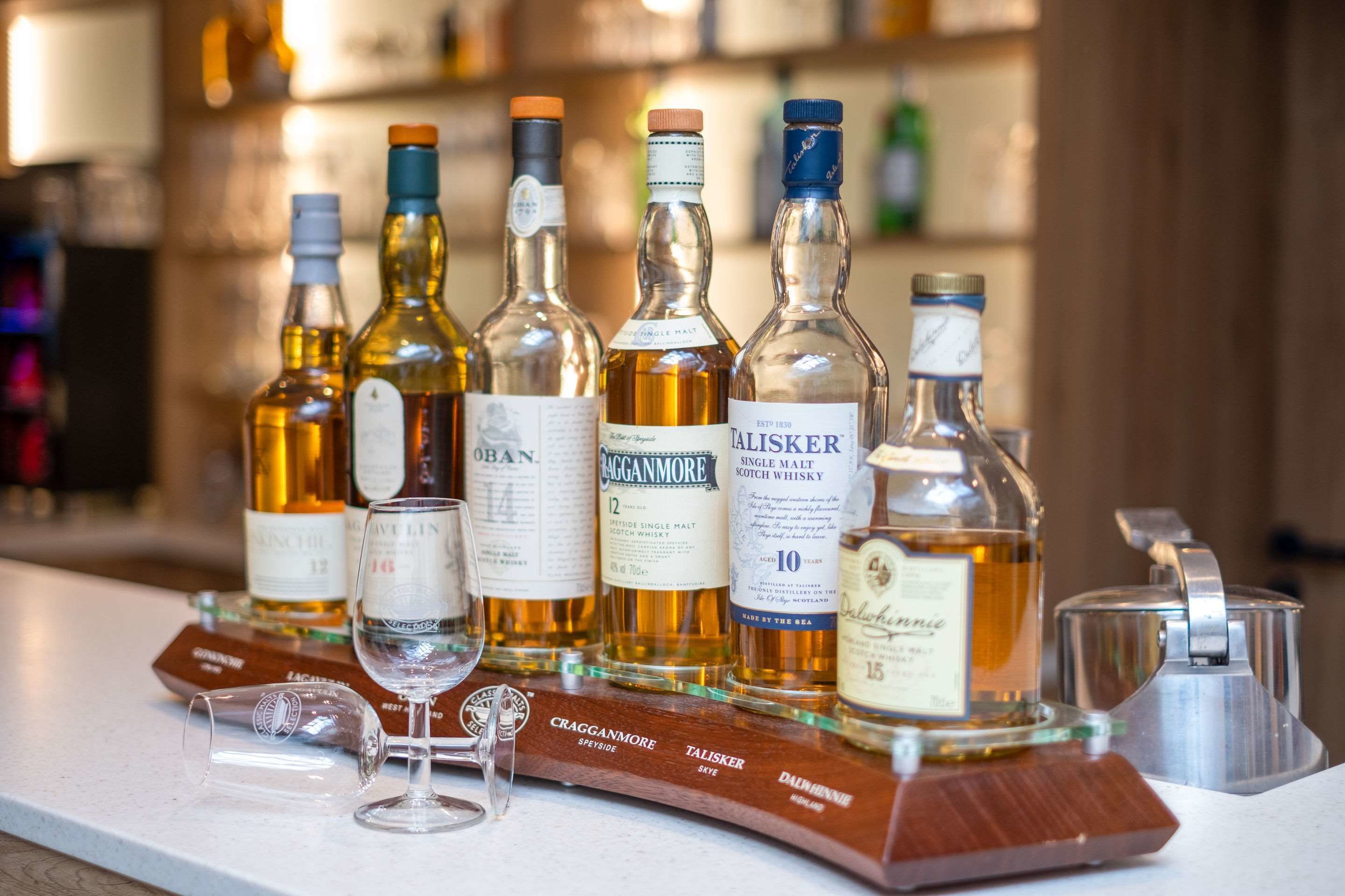 A selection of whiskey bottles on a bar counter with glasses and a wooden tray.