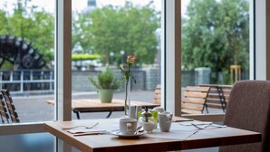 A table with a vase of flowers, cups, and a spoon, set against a window with a view of trees and a distant tower. - Quality Hotel Lippstadt (Lippstadt)