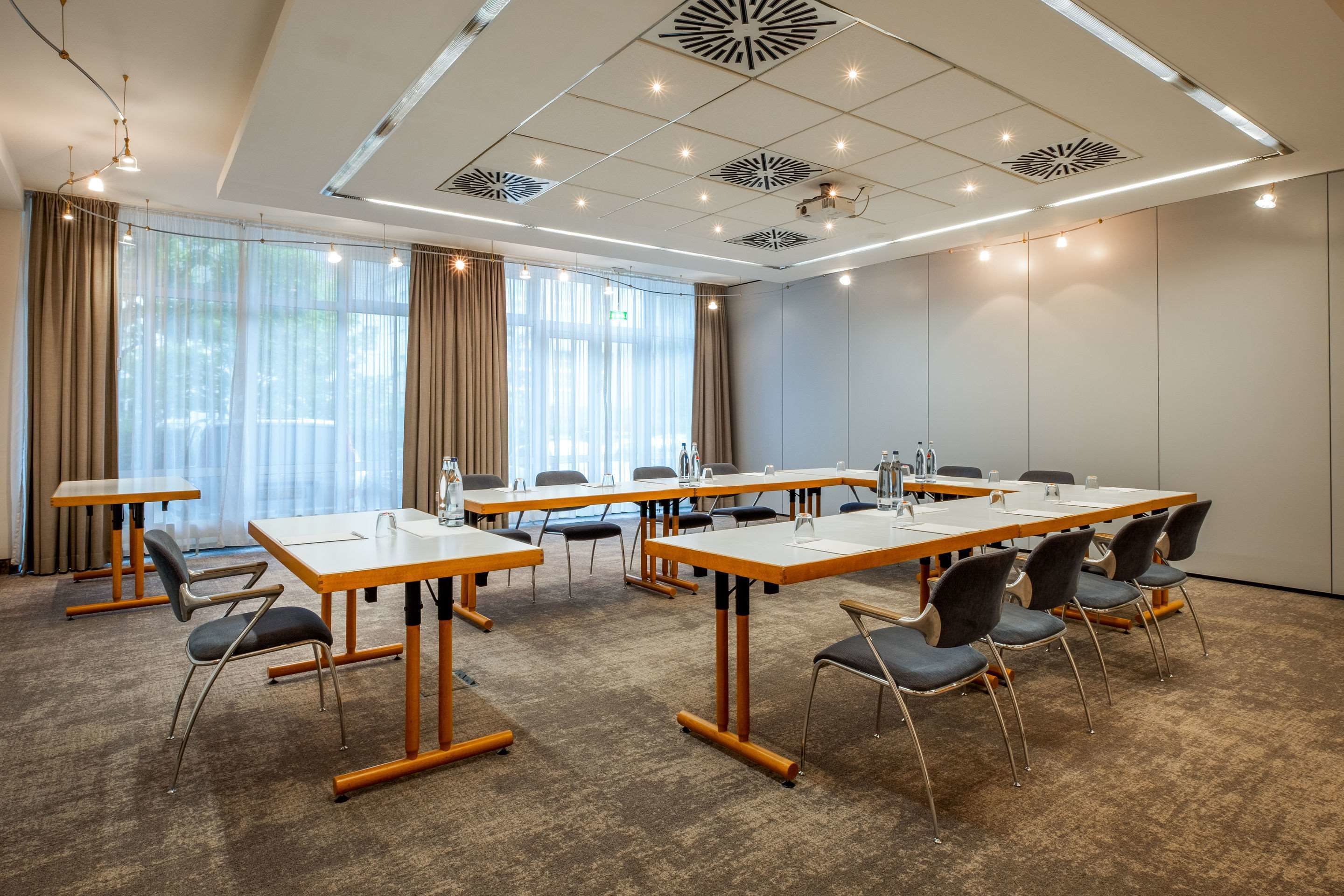 A conference room with multiple tables and chairs, large windows with curtains, and ceiling lights.