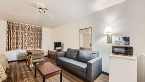 A hotel room with a gray sofa, a small wooden table, a TV, a microwave, a lamp, and a patterned curtain. - Travelodge by Wyndham Longmont (Longmont)
