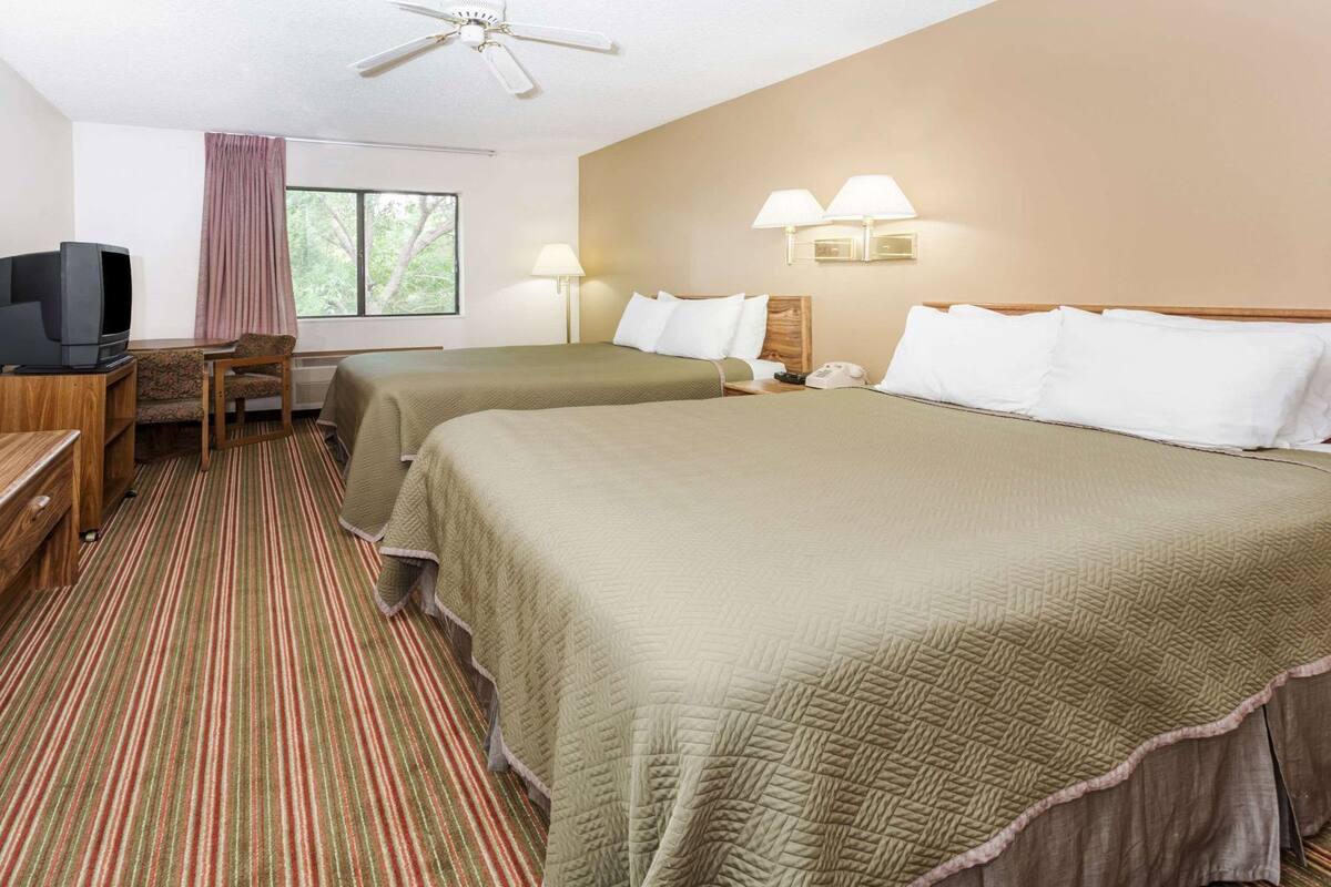 A hotel room with two beds, a desk with a television, a chair, a ceiling fan, and a window with curtains.
