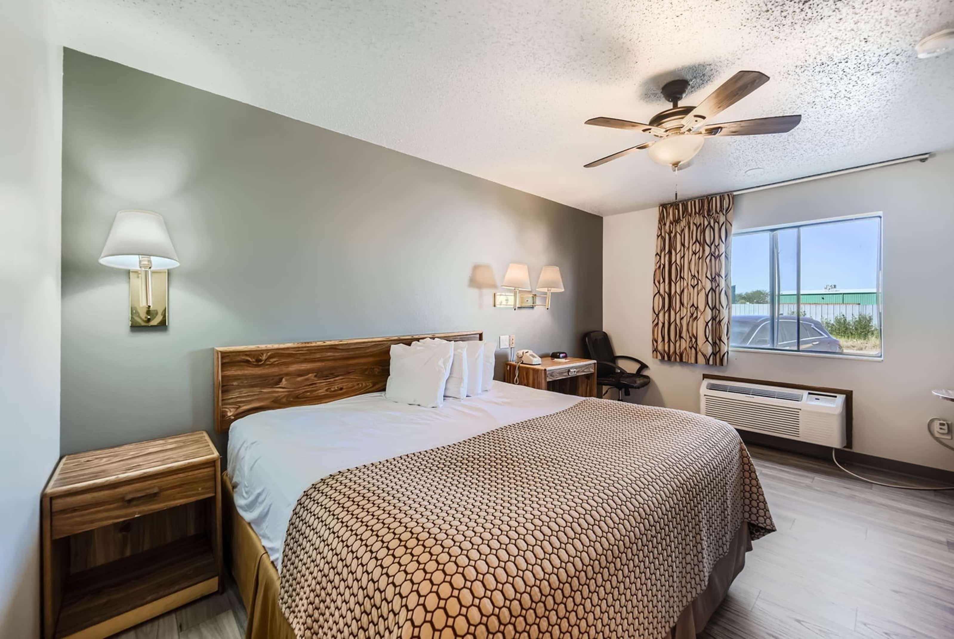 A hotel room with a bed, bedside table, lamp, wall-mounted lights, a desk, chair, window, and ceiling fan.