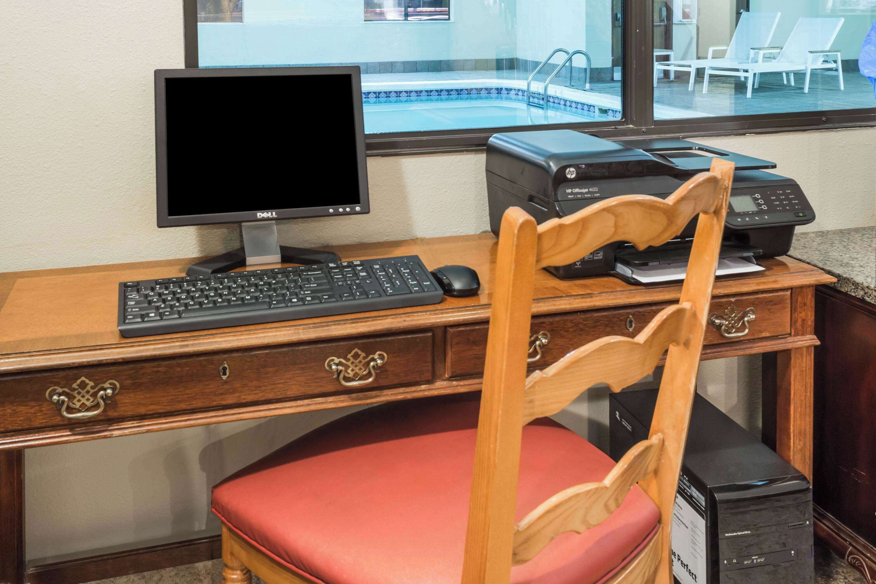 A wooden desk with a computer, keyboard, and mouse. There is a chair with a red cushion in front of the desk.