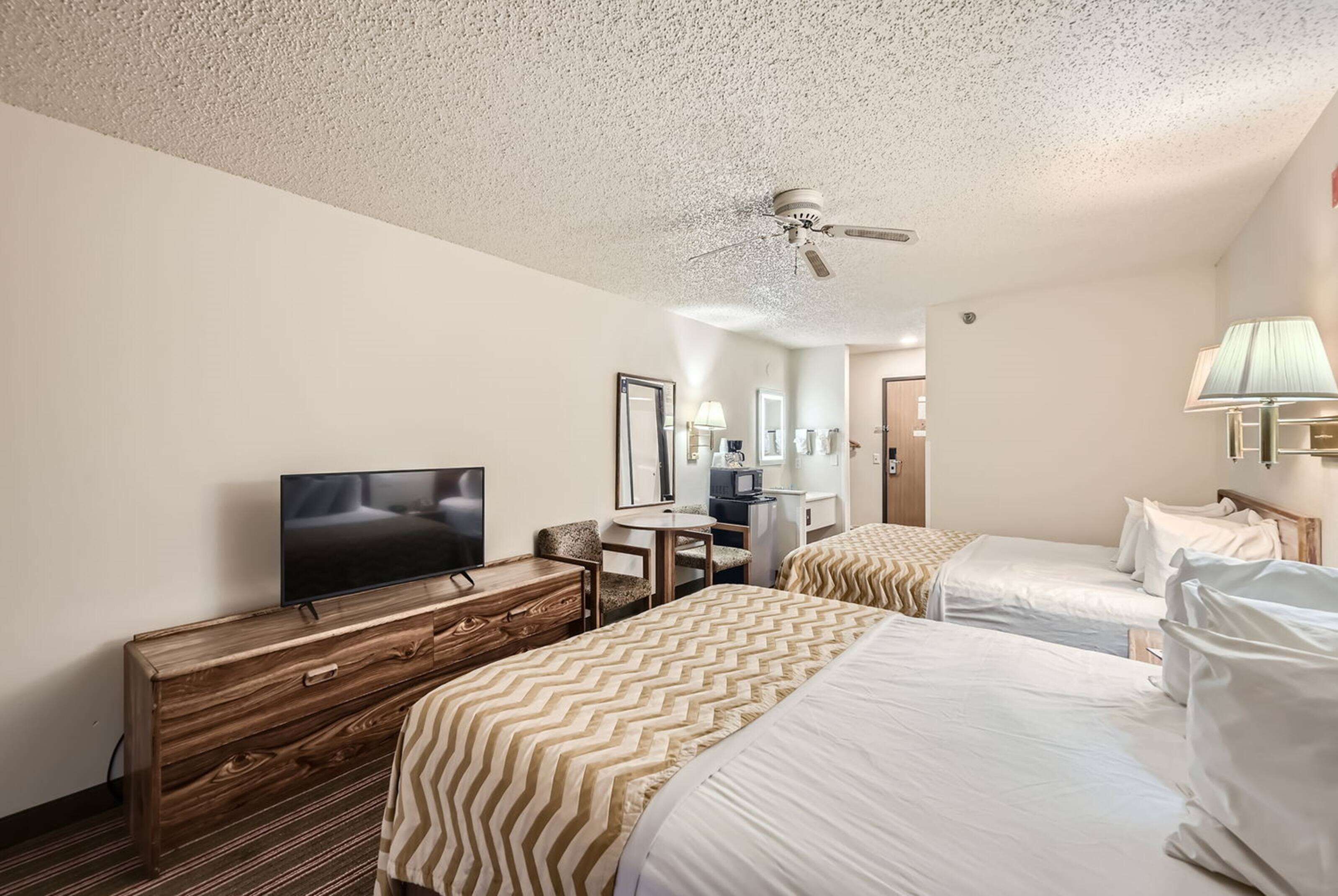 A hotel room with two beds, a TV, a wooden dresser, a desk with a chair, a ceiling fan, and a lamp.