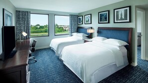A hotel room with two beds, a desk with a TV, and large windows offering a view of greenery. - Sheraton Suites Philadelphia Airport (Philadelphia)