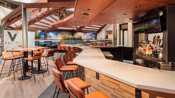 A modern bar area with wooden ceiling beams, a curved bar counter, and orange leather chairs. - Sheraton Suites Philadelphia Airport (Philadelphia)