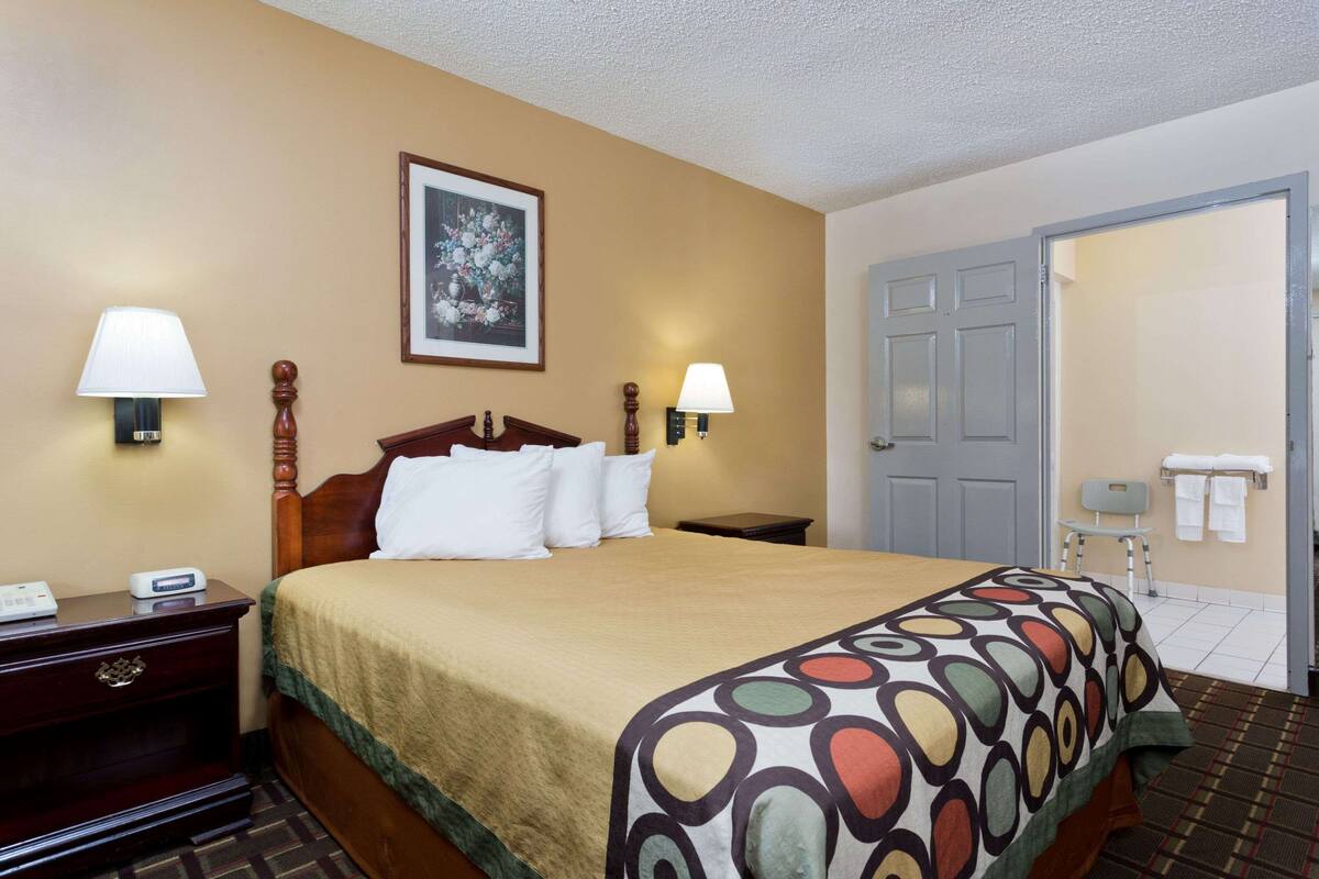 A hotel room with a bed, two bedside lamps, a nightstand, a framed picture on the wall, a door, and a chair with towels.