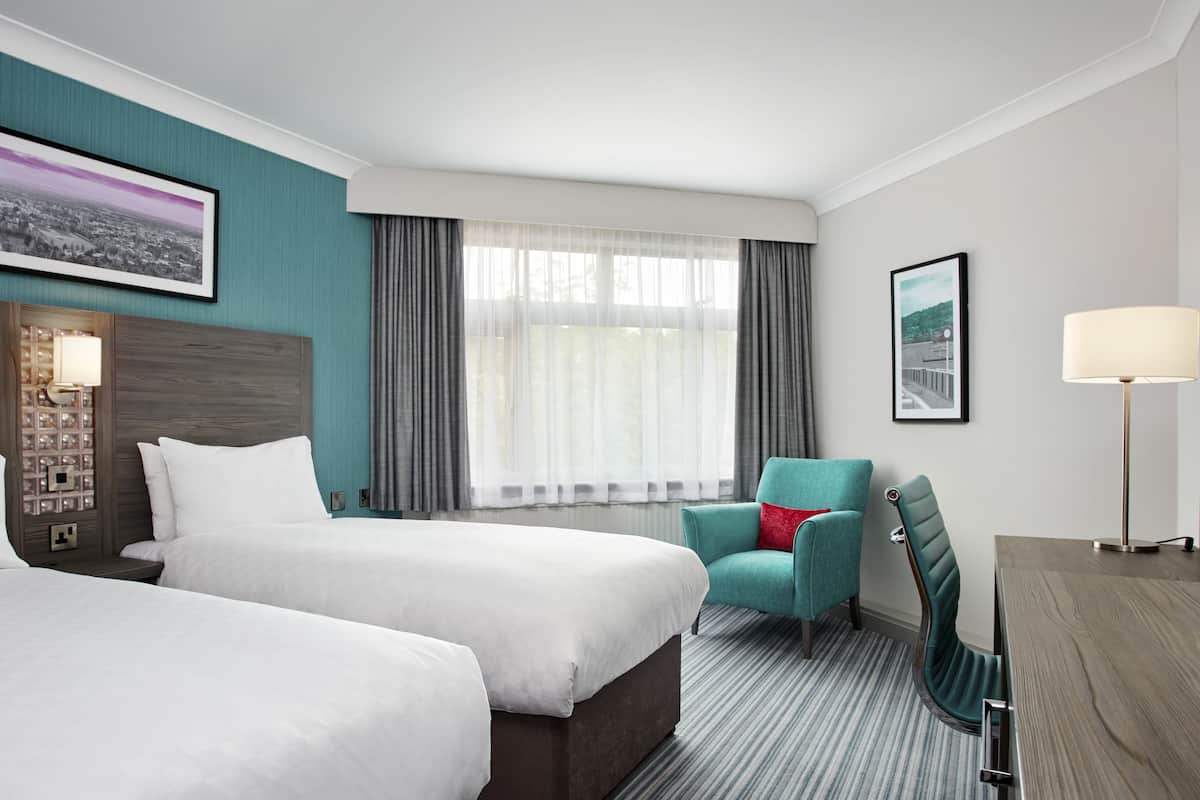 A hotel room with two beds, a desk, a chair, a lamp, and a large window with curtains.