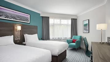 A hotel room with two beds, a desk, a chair, a lamp, and a large window with curtains.