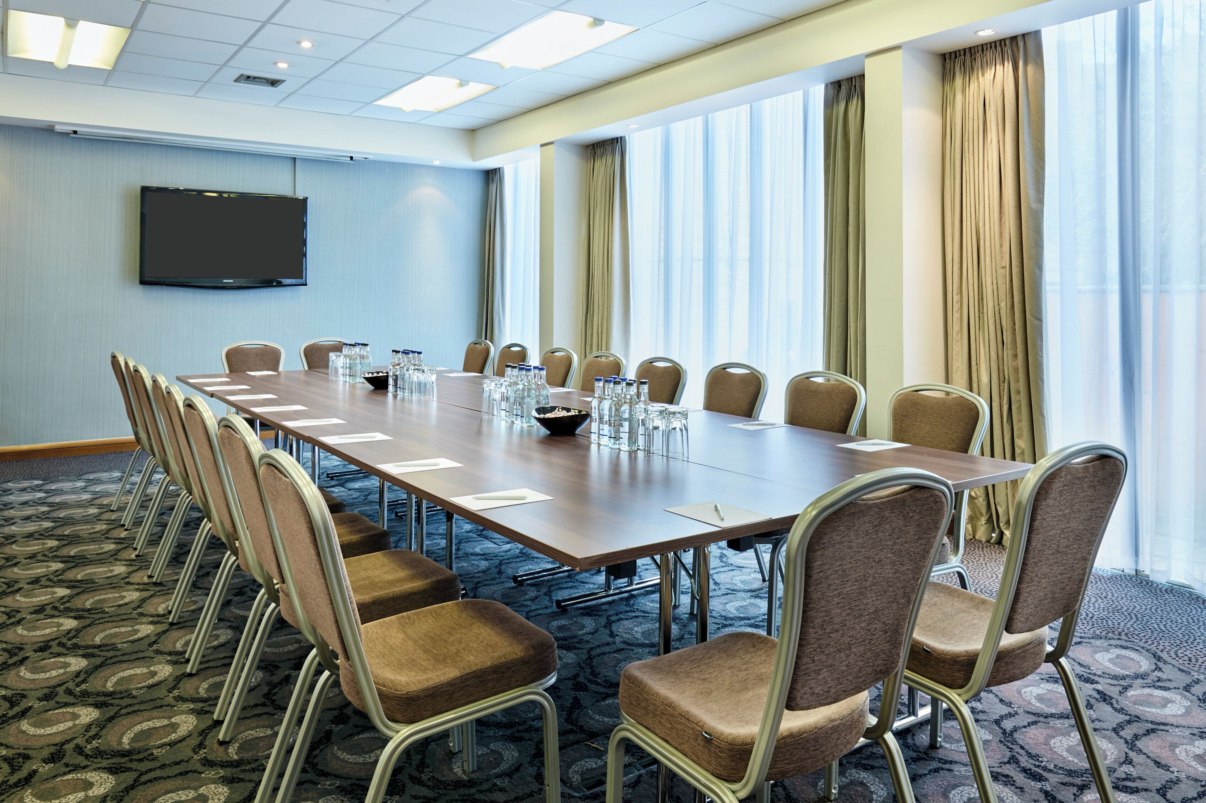A conference room with a long table, chairs, a flat-screen TV, and a large window with curtains.