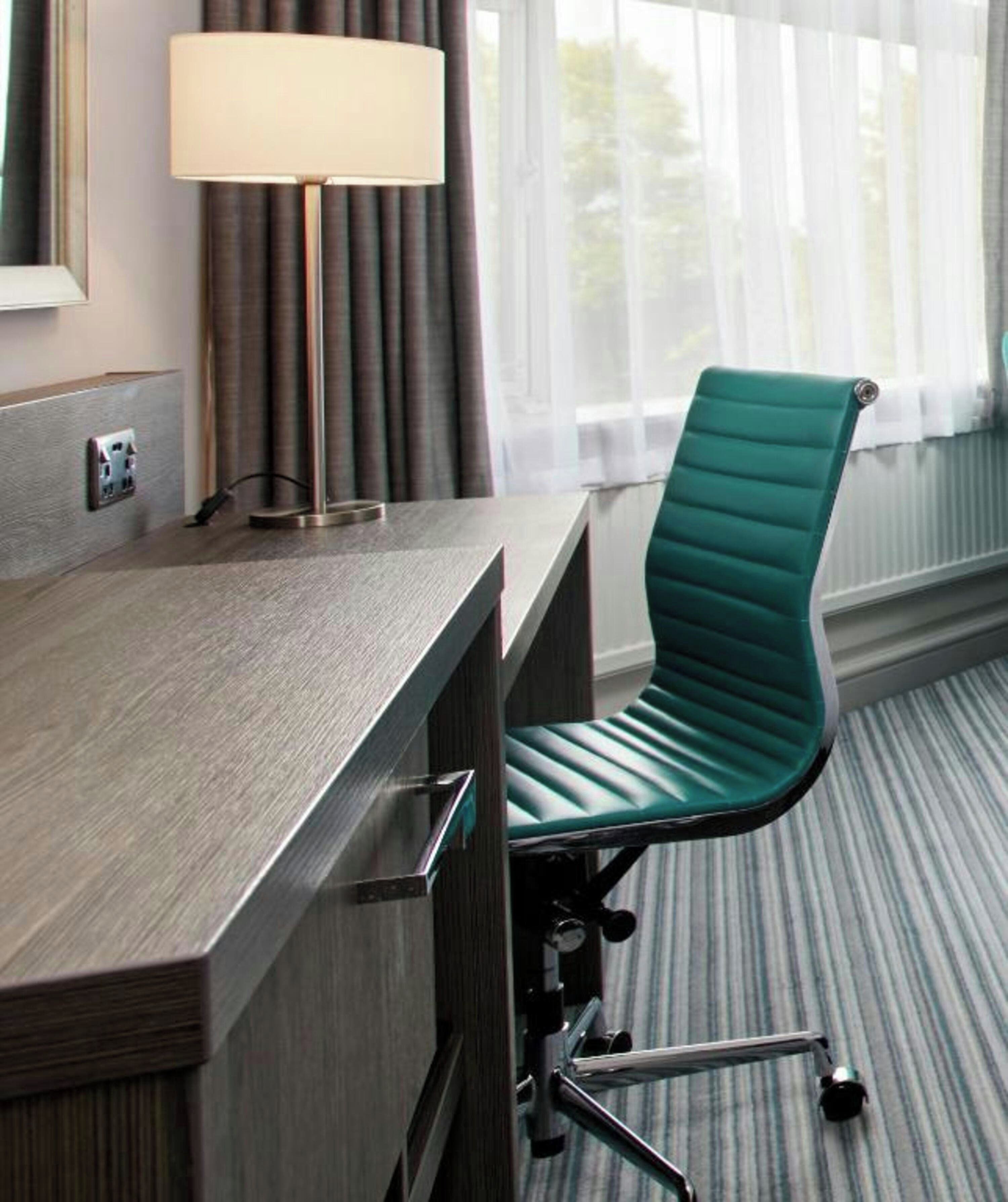 A hotel room with a desk, chair, lamp, and window with curtains.