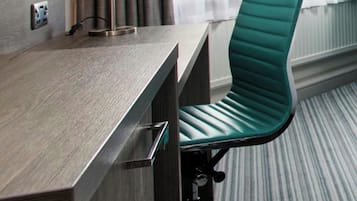 A hotel room with a desk, chair, lamp, and window with curtains.