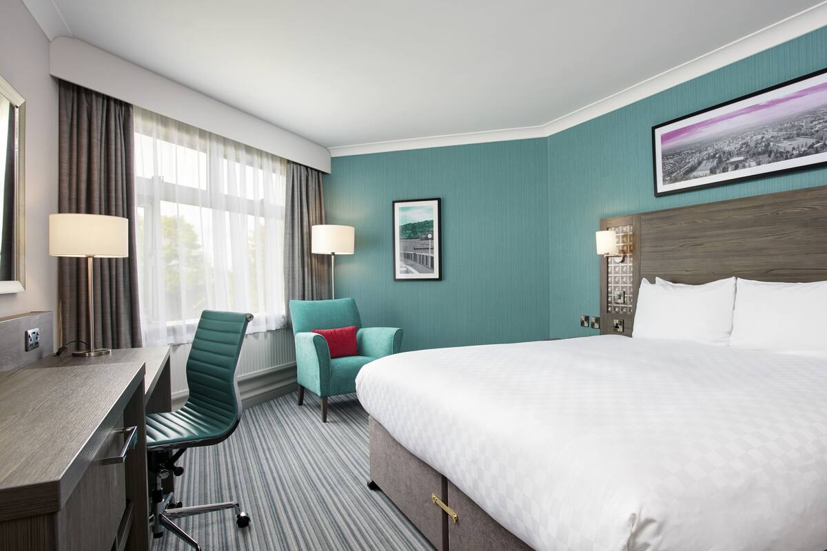 A hotel room with a large bed, a desk with a chair, a window with curtains, and a framed picture on the wall.