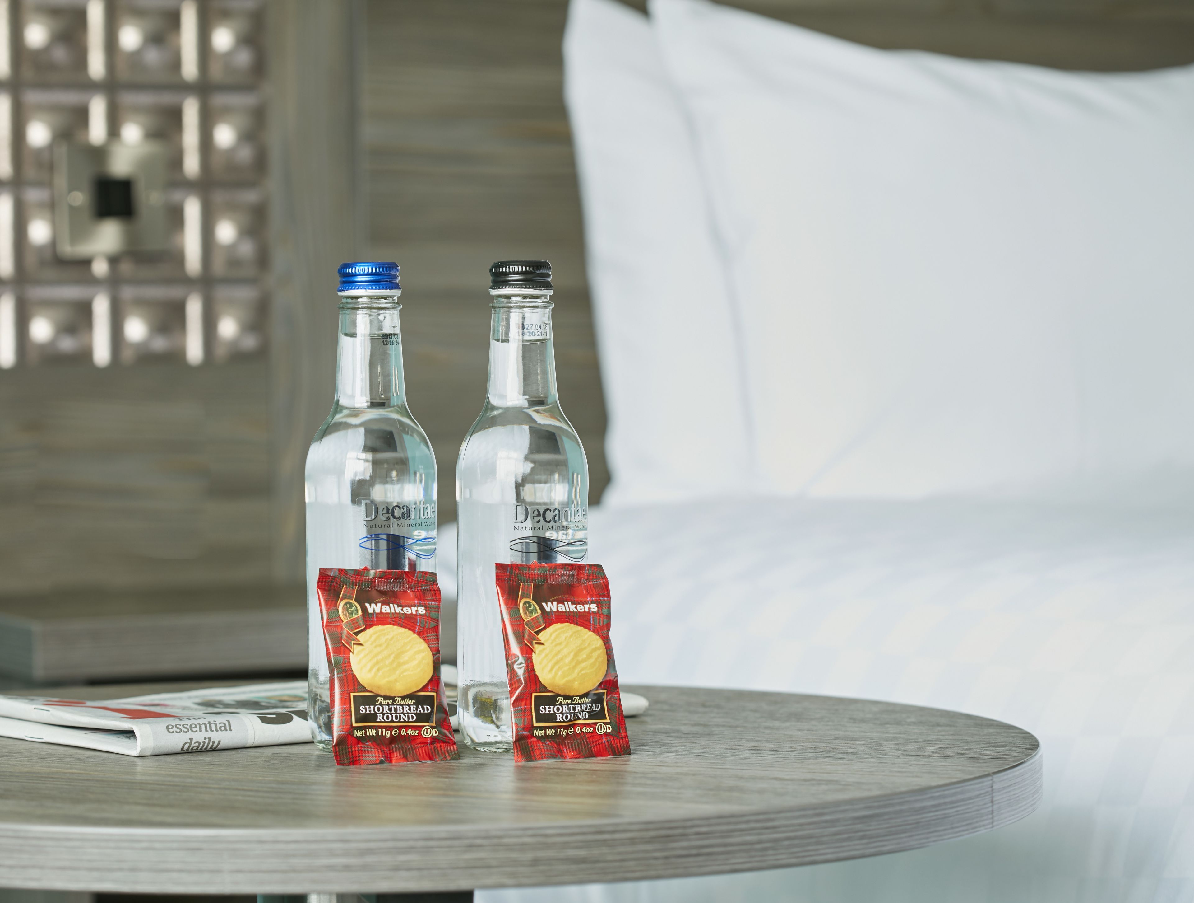 Zwei Flaschen Decanta Mineralwasser und zwei Packungen Walkers Shortbread Rounds auf einem runden Tisch in einem Hotelzimmer.