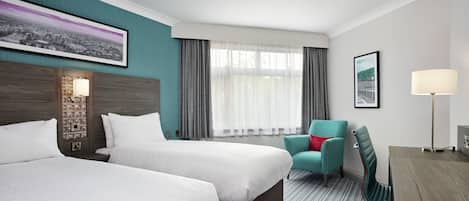 A hotel room with two beds, a chair, a desk, a lamp, and a window with curtains.