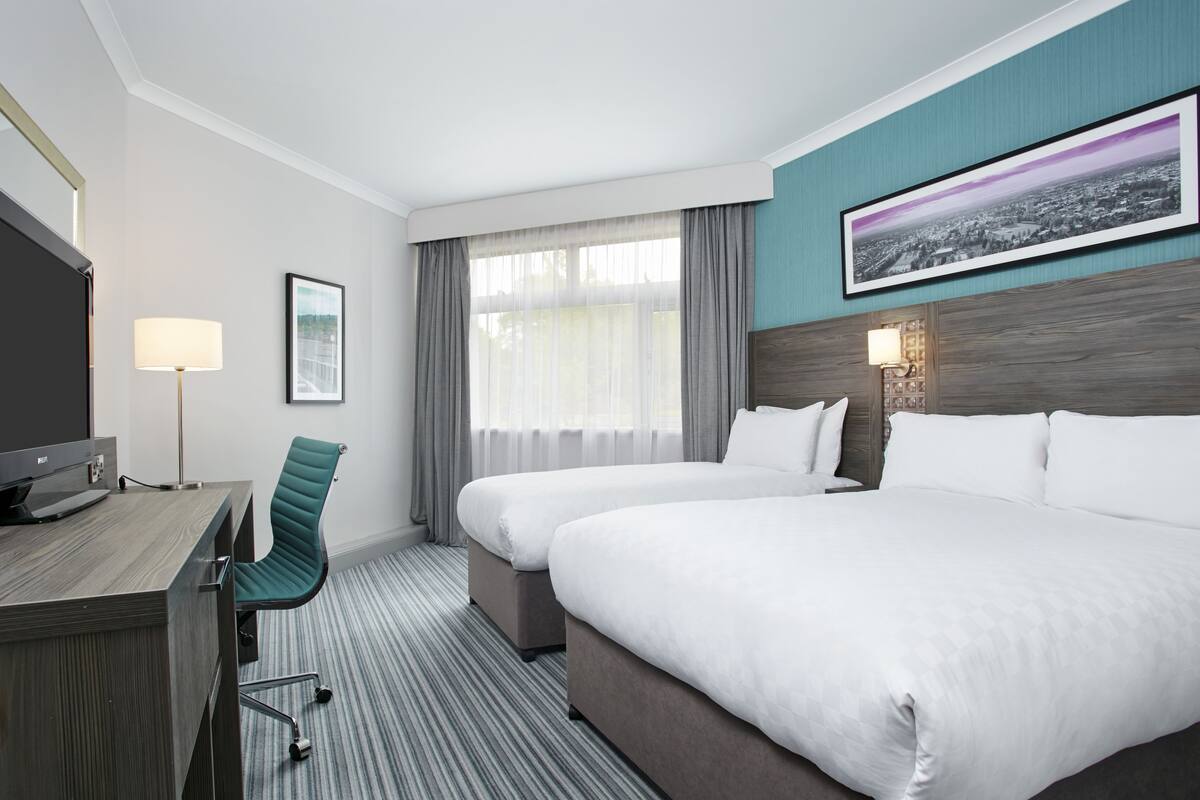 A hotel room with two beds, a desk, a TV, a chair, and a window with curtains.
