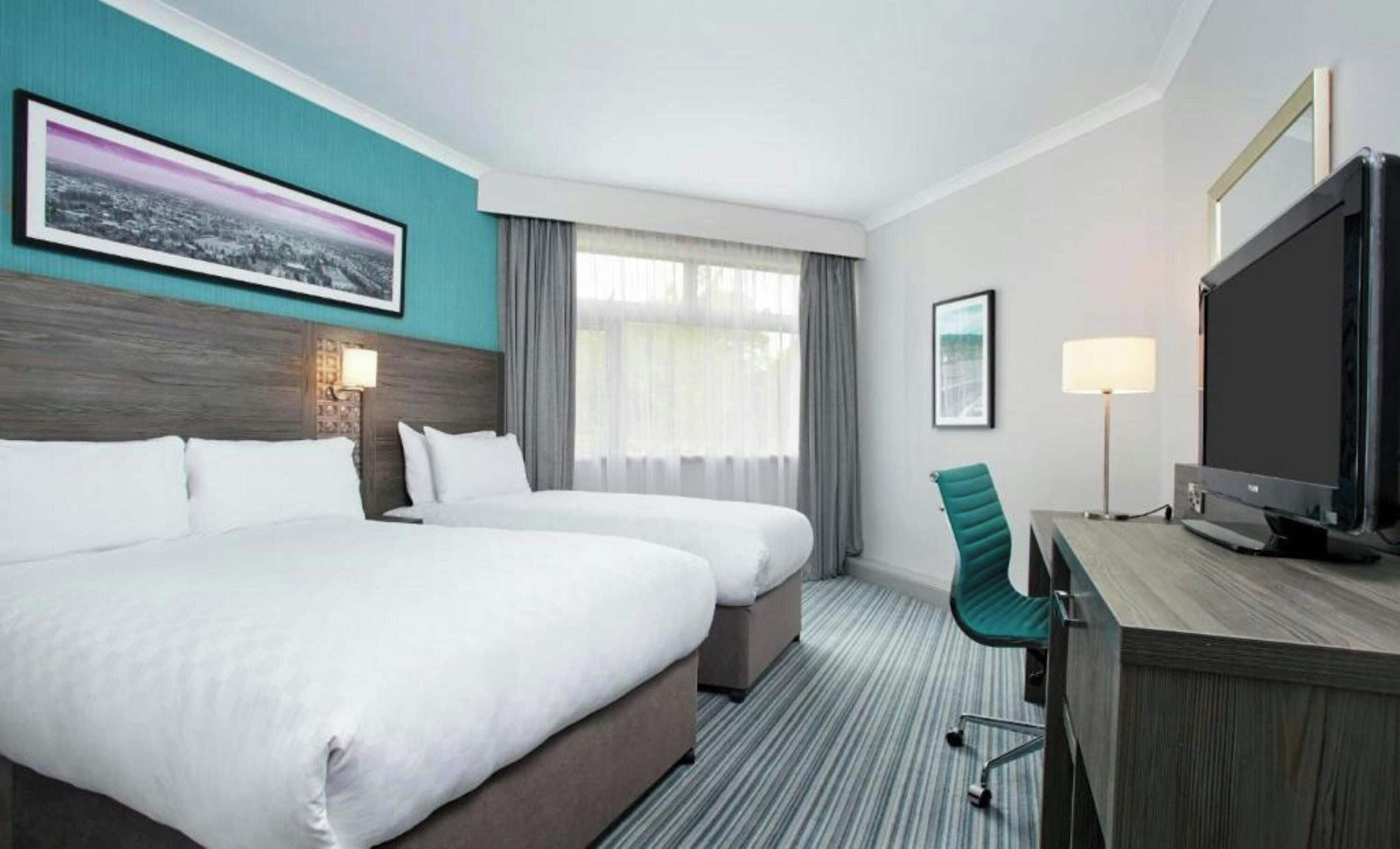 A hotel room with two beds, a desk with a television, a chair, and a window with curtains.