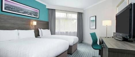 A hotel room with two beds, a desk with a television, a chair, and a window with curtains.