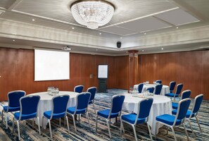 A conference room with round tables, blue chairs, a projector screen, and a large chandelier. - Leonardo Hotel Cheltenham (Cheltenham)