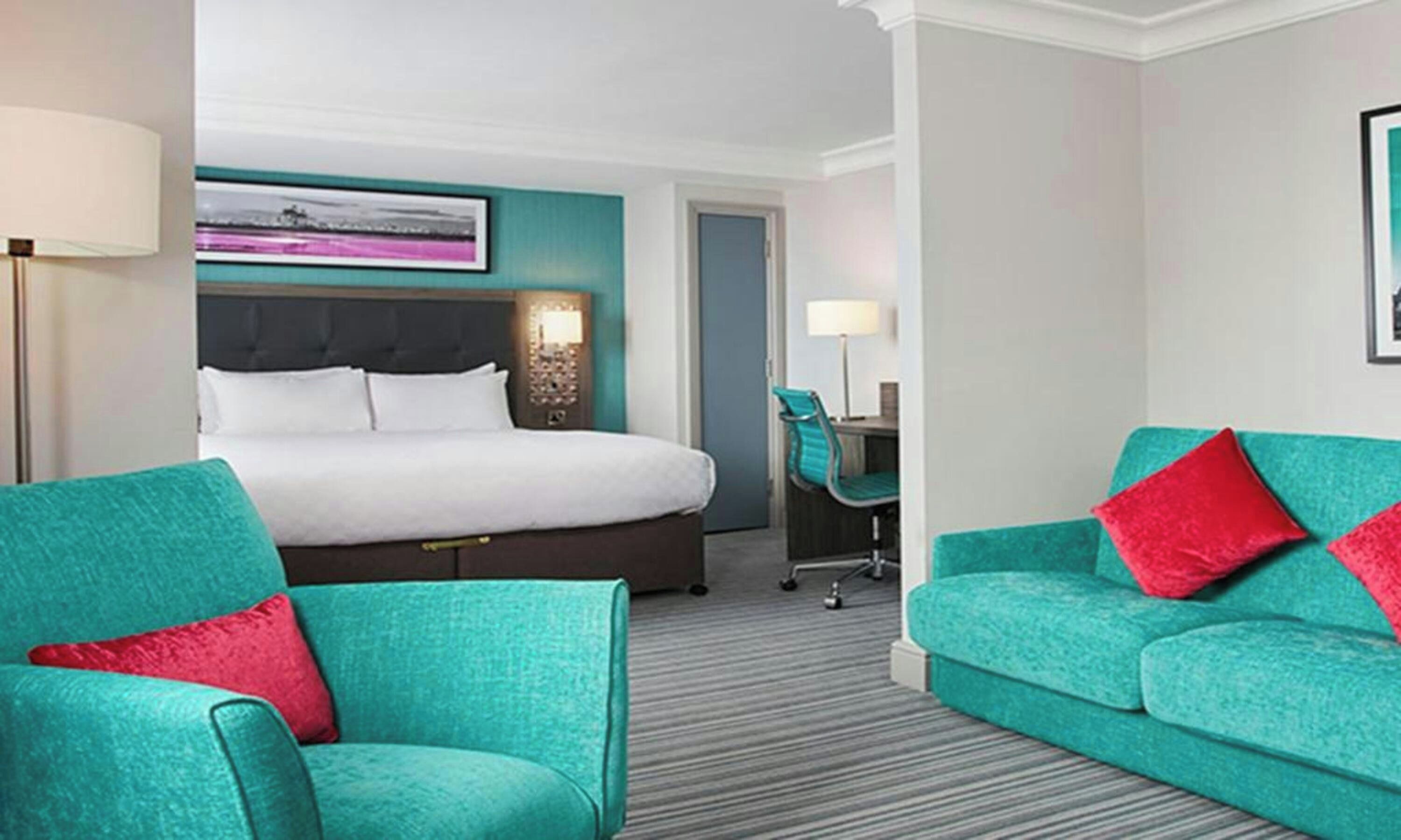 A hotel room with a bed, two teal armchairs, a desk, and a chair.