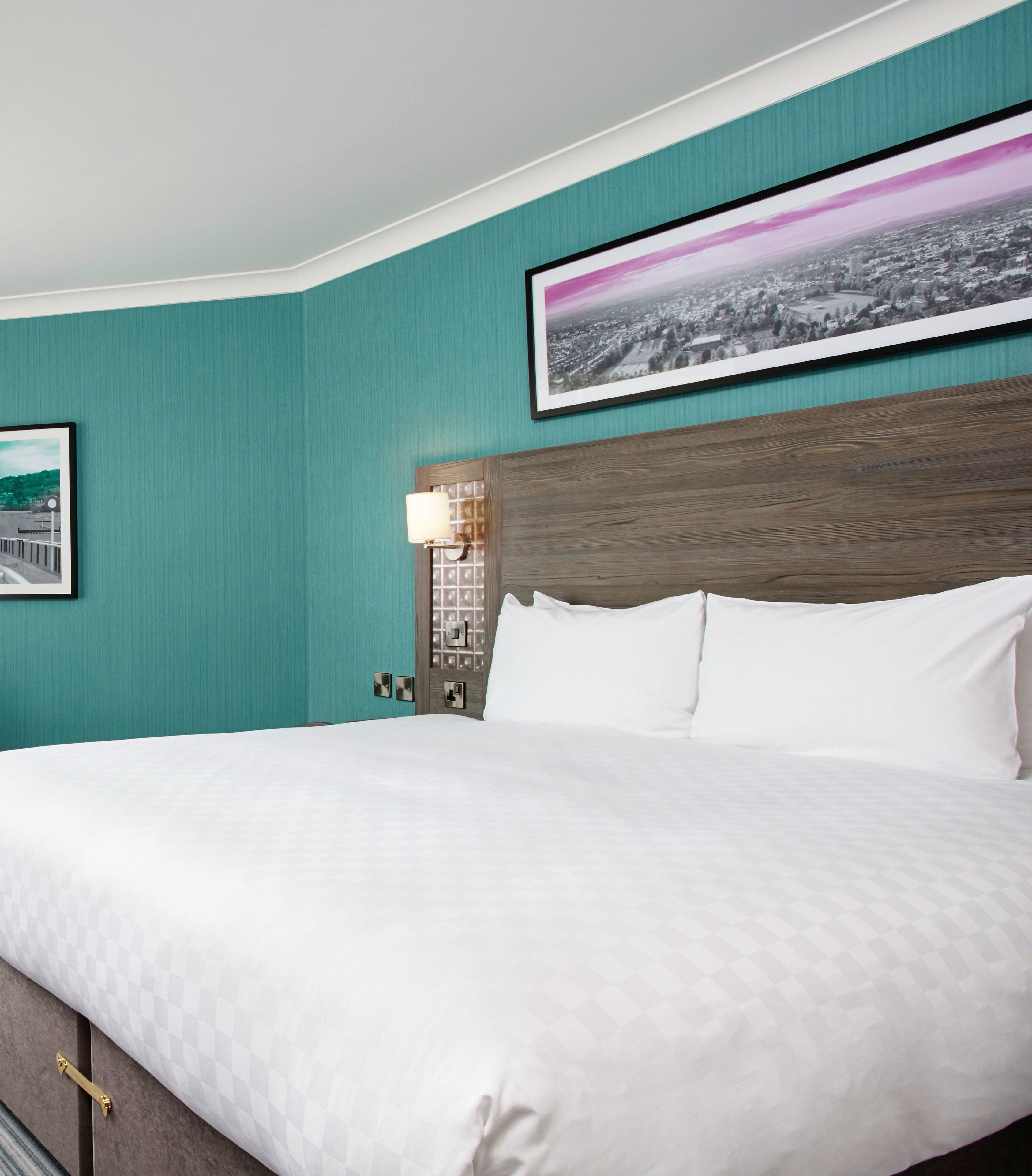 A neatly made bed in a hotel room with teal walls, a wooden headboard, and a cityscape picture above it.
