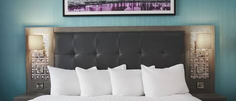 Una habitación de hotel con una cama grande, una obra de arte con un paisaje urbano sobre la cama, y dos lámparas de noche.