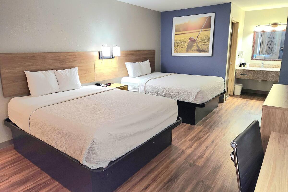 A hotel room with two beds, a wooden headboard, a nightstand with a lamp, a painting on the wall, and a bathroom with a mirror and sink.