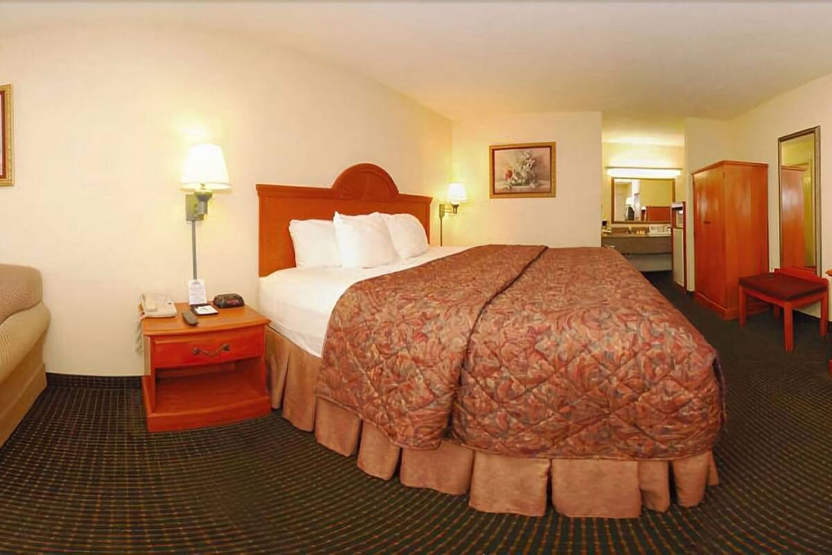 A hotel room with a bed, bedside table with a lamp, a chair, a wardrobe, and a mirror.